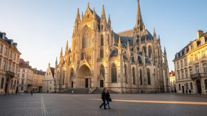 Vue réaliste de la cathédrale Saint-Étienne de Metz depuis la place d’Armes, avec sa façade gothique en pierre de Jaumont éclairée par une lumière dorée de fin de matinée et quelques passants discrets.